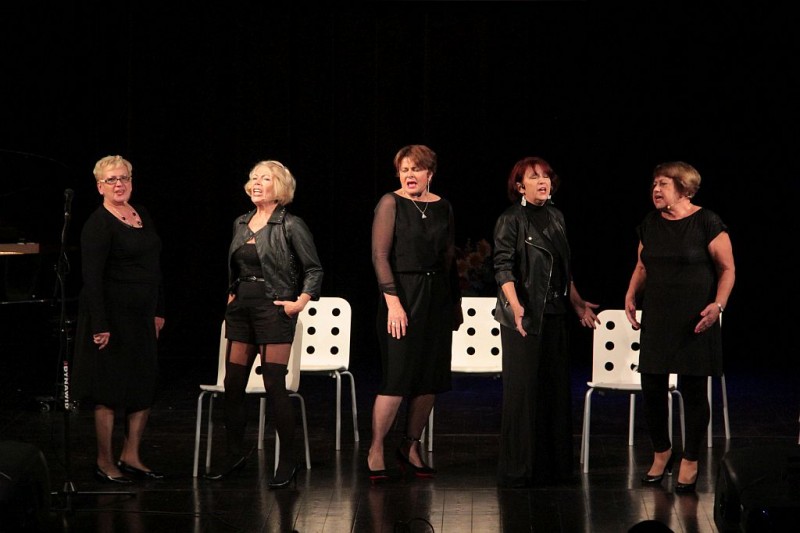 Na scenie pięć kobiet w czarnych sukienkach. Za nimi białe krzesła