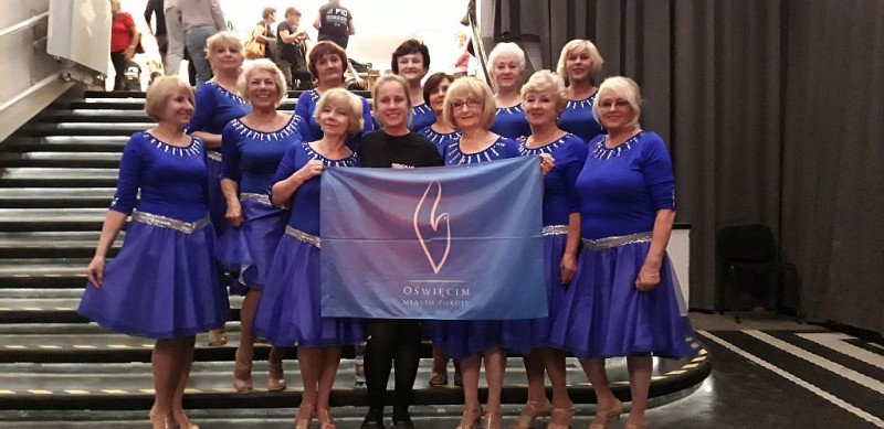 Kobiety w niebieskich sukienkach stoją na schodach, jedna z nich trzyma flagę miasta Oświęcim