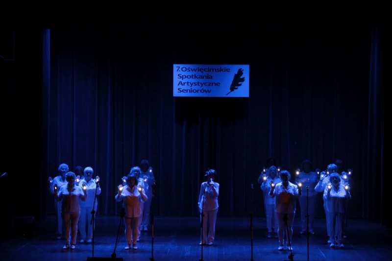 Artyści wyśsępują na scenie przy zgaszonych światłach. W rękach trzymają małe latarki.