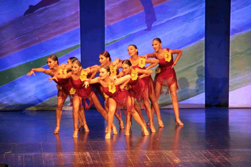 Zespół dziewczęcy tańczy na scenie w czerwonych sukienkach z przypiętym żółtym kwiatem