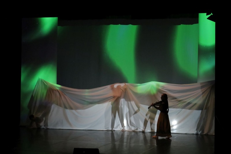 Na scenie kobieta grająca na skrzypcach i trzy osoby przykryte płachtą