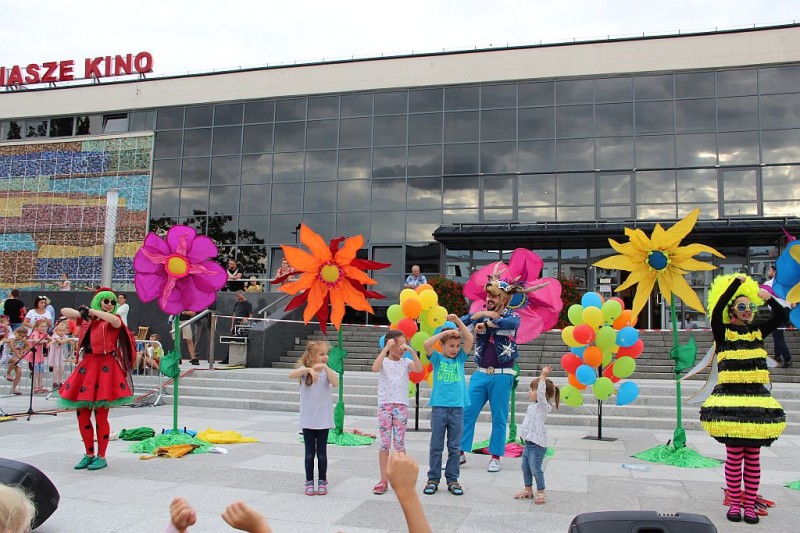 Dzieci tańczą z postaciami z bajek. Za nimi stoją duże ręcznie wykonane kwiaty