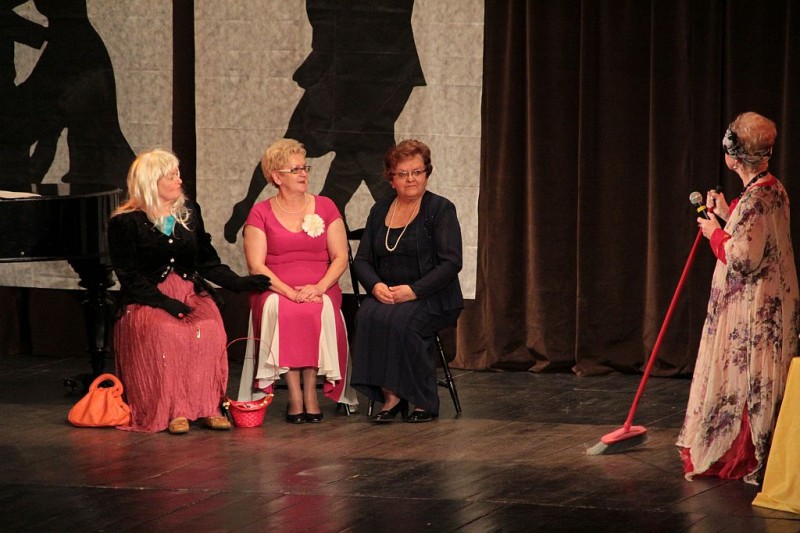Trzy kobiety siedzą na krzesłach. Obok stoi kobieta z miotłą w ręce.