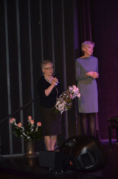 Dwie kobiety stoją na scenie. Jedna przemawia a w ręce trzyma bukiet kwiatów.
