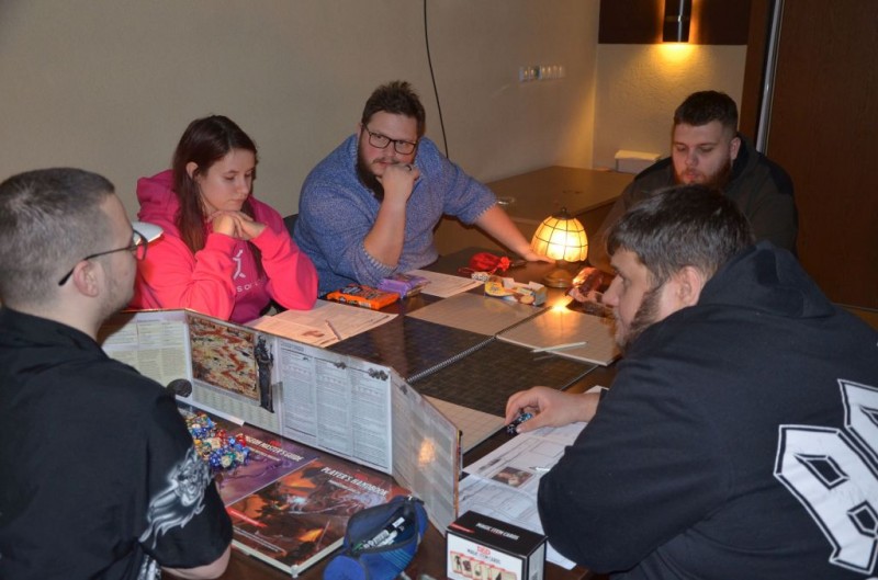 Gracze siedzą przy stole z planszami do gry, na środki stoi lampka nocna