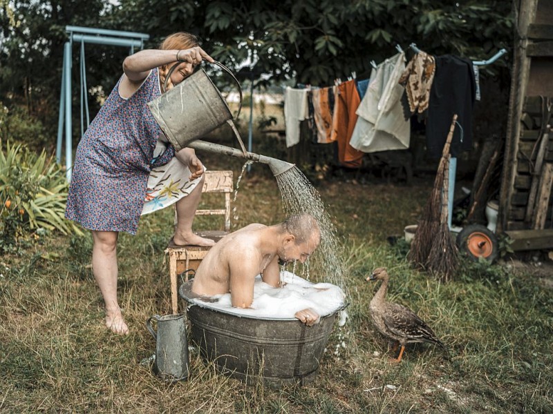 Mężczyzna siedzi w bali z woda i pianą. Nad nim Stoi kobieta i polewa go wodą z dużej konewki.