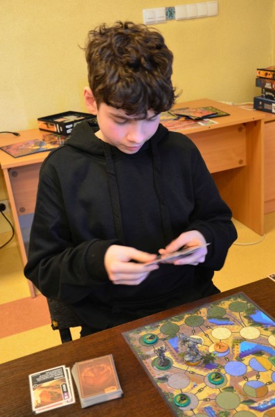 Młody chłopak siedzi nad plansza a w rękach trzyma karty do gry