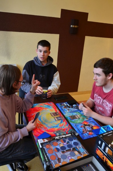 Trzy osoby siedzą przy stole, przed nimi rozłożone są plansze do gry