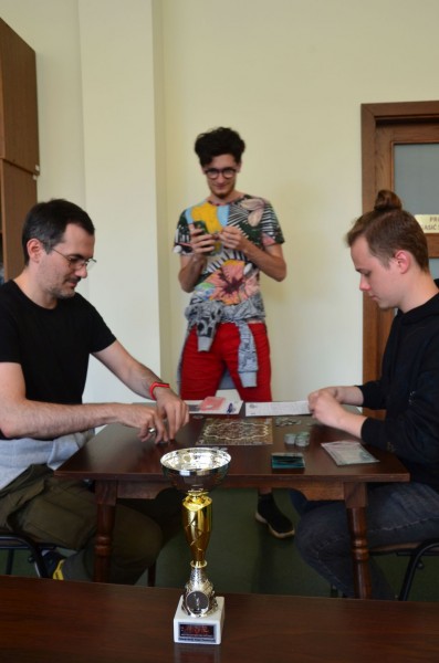 Dwóch mężczyzn gra przy stoliku, trzeci stoi obok nich